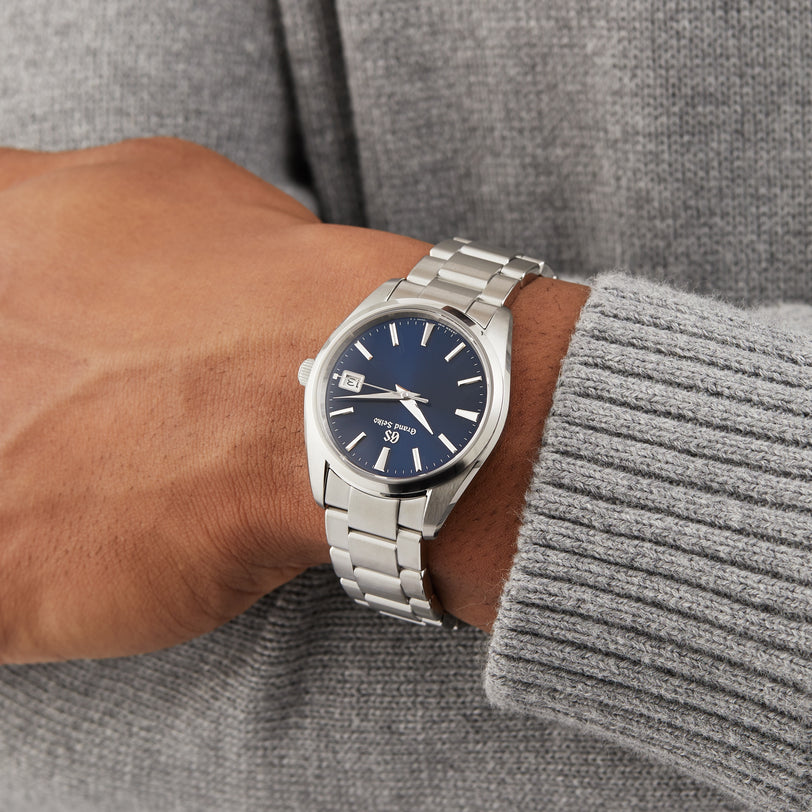 A person wearing a Grand Seiko Quartz Heritage stainless steel watch with a blue face and date display, paired with a light gray knit sweater.