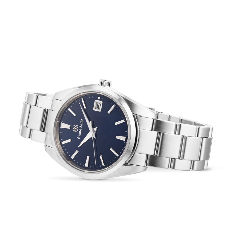The Grand Seiko Quartz Heritage is a stainless steel watch with a blue dial, silver hour markers, a date window at 3 o’clock, and a stylish metal link bracelet.
