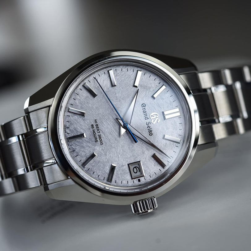 A close-up of the Grand Seiko Hi-Beat Heritage wristwatch with a textured silver dial, blue second hand, 3 o’clock date display, and Ever-Brilliant Steel bracelet highlights its precise Hi-Beat 36000 movement.