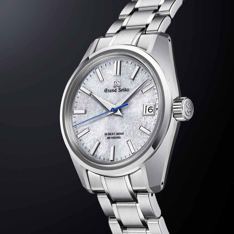 The Grand Seiko Hi-Beat Heritage wristwatch showcases a textured white dial, silver markers, blue second hand, date at 3 o’clock, and an Ever-Brilliant Steel bracelet set against a dark gradient background.