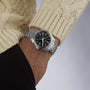 A person in a cream knit sweater and black top is wearing the Grand Seiko Hi-Beat GMT Heritage watch featuring a silver case, black dial, metal band, and precise automatic movement.