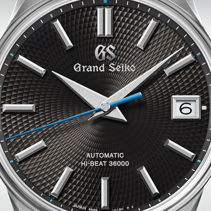 Close-up of the Grand Seiko Hi-Beat Heritage wristwatch featuring a textured black dial, silver hour markers, a blue seconds hand, and a date window showing the number 6.