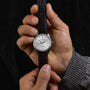 A person holds a Grand Seiko Manual Hi-beat 80 Hours Evolution 9 wristwatch featuring a textured white birch dial, silver case, and black leather strap against a black background.