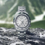 The Grand Seiko Hi-Beat GMT Sport wristwatch, featuring a textured silver dial and green bezel, is displayed on a rock with blurred mountains in the background.