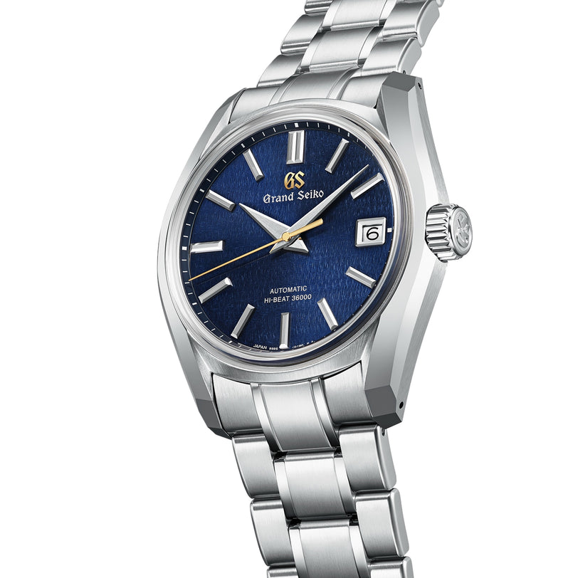 The Grand Seiko Hi-Beat Heritage is a stainless steel wristwatch with a blue textured dial, silver hour markers, date window at 3 o’clock, Zaratsu polishing, and a matching silver bracelet.