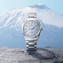The Grand Seiko Hi-Beat 80 Hours Evolution 9 wristwatch with a textured silver face is displayed on a rock, set before the blurred backdrop of a snow-capped mountain landscape.