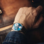 A wrist wearing the MB&F Legacy Machine LM101 Steel Blue luxury watch rests on a brown leather bag, with the person dressed in a denim jacket.