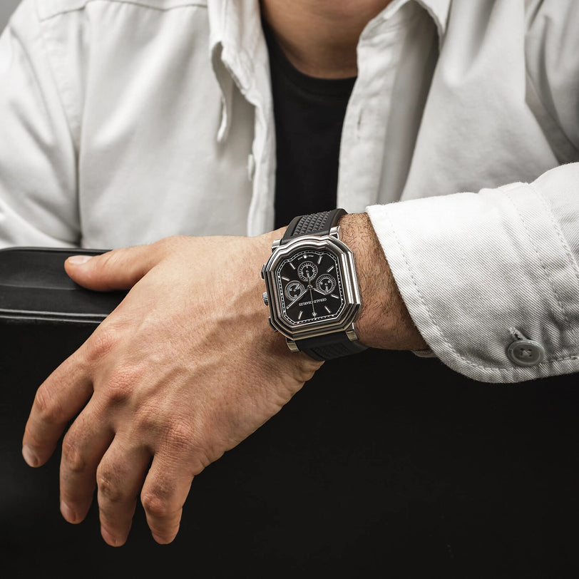 A person wearing a white shirt and black t-shirt showcases the Gerald Charles Maestro 3.0 Chronograph, a black and silver stainless steel watch, on their left wrist while resting their arm on a black surface.
