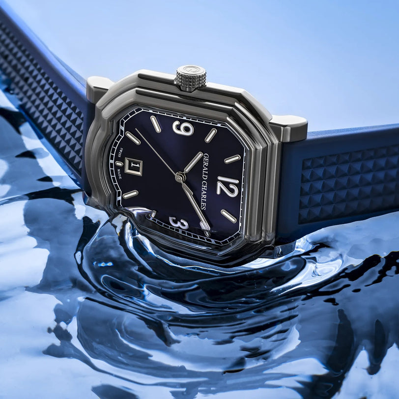 The Gerald Charles Maestro GC Sport watch, featuring a blue vulcanized rubber strap and silver case, rests partially submerged in rippling water and displays the time 1:00.