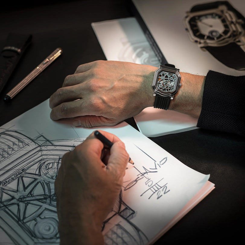 A person sketches a watch design, wearing the Gerald Charles Maestro 8.0 Squelette skeleton dial wristwatch, with pen and sketches in view.