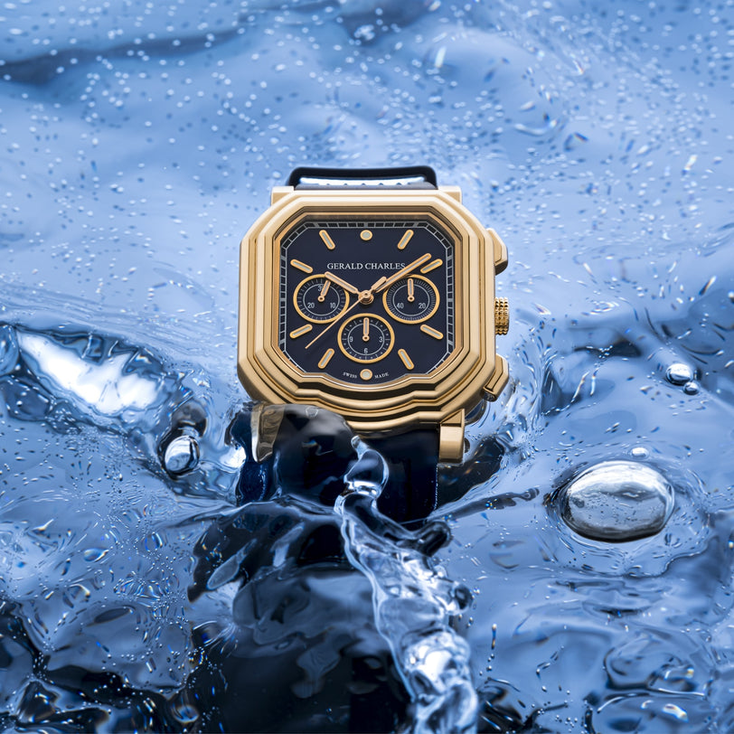 The Gerald Charles Maestro 3.0 Chronograph, a gold luxury watch with a black face, is partially submerged in shimmering blue water with splashes.
