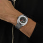 Wearing a DOXA Sub 300 Sharkhunter—silver divers watch with black dial and green accents, unidirectional bezel—a person stands against a black background in a black zippered jacket.