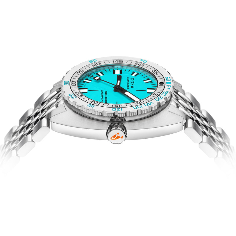 The DOXA Sub 300T Aquamarine is a silver divers watch featuring a vibrant aquamarine dial, black hour markers, and a stainless steel bracelet, highlighted from a low angle with its crown, case side, and helium release valve.