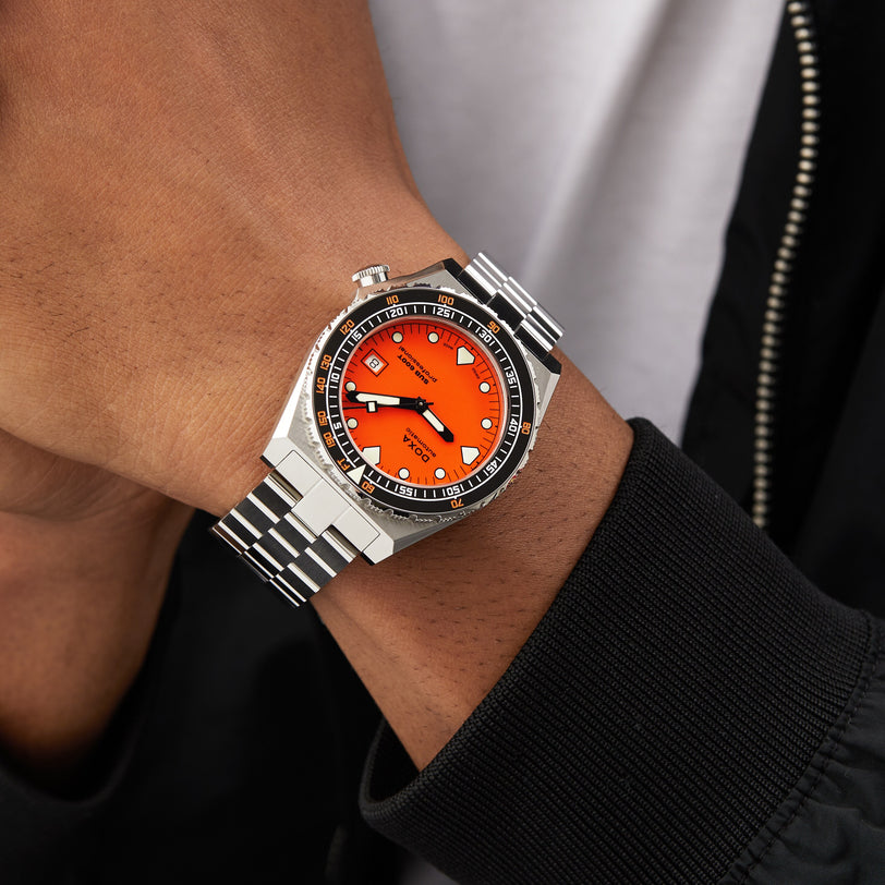 A person wearing a DOXA Sub 600T Professional dive watch with a silver bracelet, orange dial, and black bezel, dressed in a black jacket and white shirt.