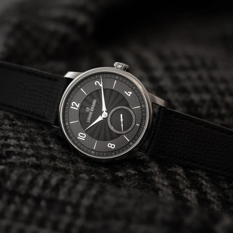 A Louis Erard Petite Seconde Guilloché Anthracite Black wristwatch with a textured dial is displayed on a dark, patterned fabric background.