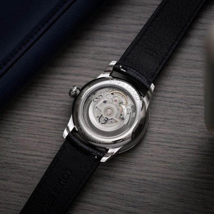 A close-up of the Louis Erard Petite Seconde Guilloché Anthracite Black’s transparent case back reveals its mechanical movement, set on a wooden surface.