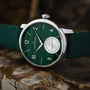 The Louis Erard Petite Seconde Malachite watch features a green dial with silver details and a round subdial, displayed on a brown stone surface.
