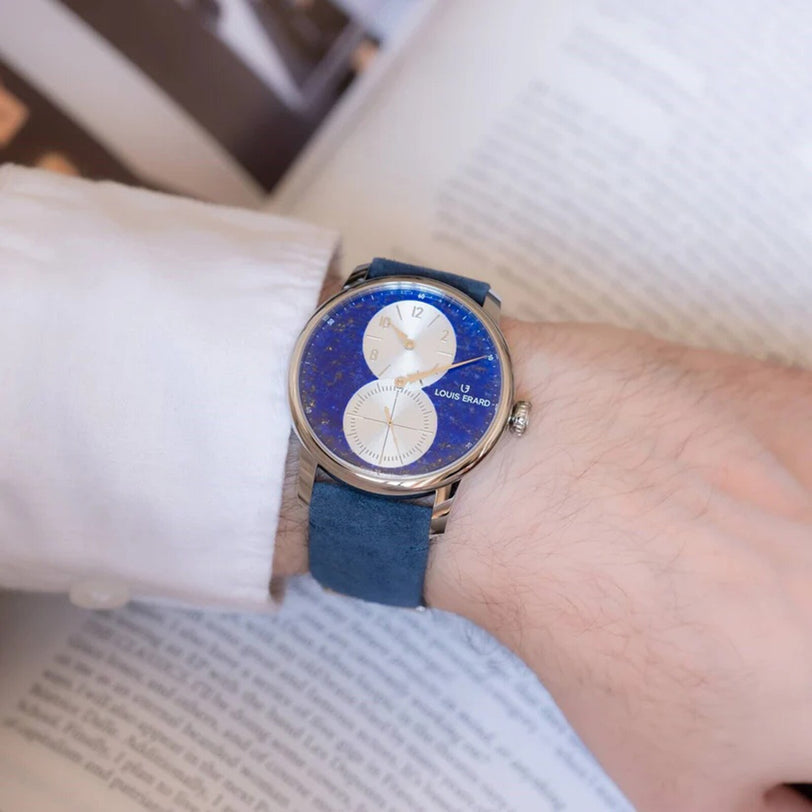 A person wearing a Louis Erard Régulateur Lapis-Lazuli wristwatch with a white dial, resting on an open book.