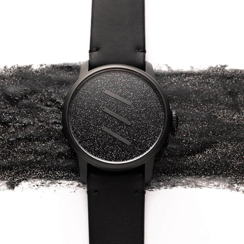 The Louis Erard Le Régulateur Louis Erard x Olivier Mosset is a black wristwatch featuring a textured, glittery face with no numbers and comes on a matching black leather strap.