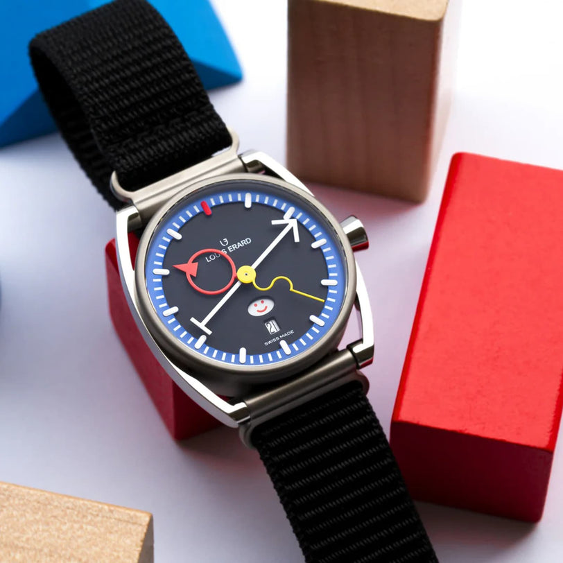 The Louis Erard La Semaine Louis Erard x Alain Silberstein wristwatch features a playful black dial, colorful hands, and a black strap, shown surrounded by wooden blocks.