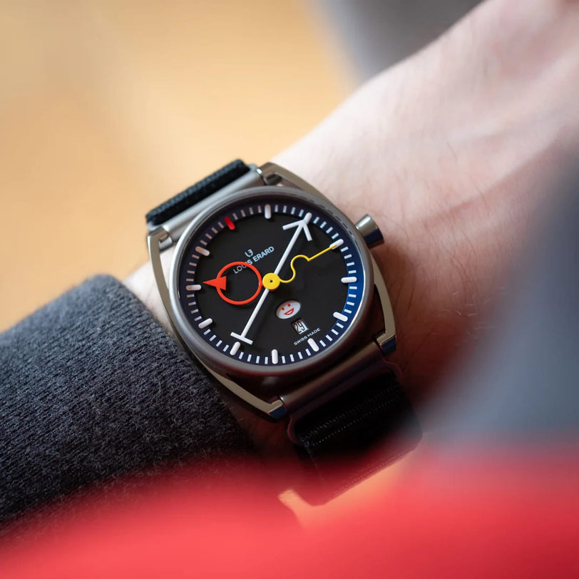 The Louis Erard La Semaine Louis Erard x Alain Silberstein wristwatch, featuring red, yellow, and white hands and a black strap, is shown on a persons wrist.