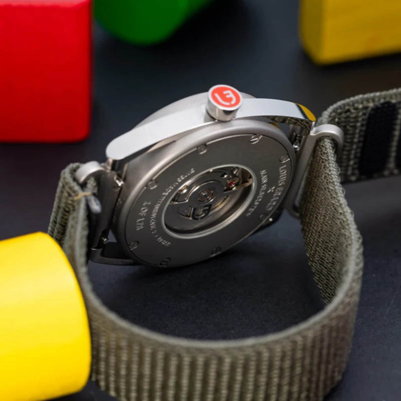 Close-up of the Louis Erard Le Régulateur x Alain Silberstein Khaki wristwatch back with a grey fabric strap, surrounded by colorful wooden blocks.