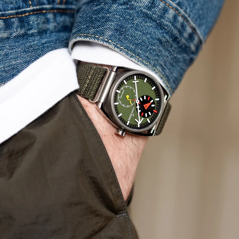 Close-up of the Le Régulateur Louis Erard x Alain Silberstein Khaki wristwatch with a green strap, worn on a wrist with hand in pocket and denim jacket.