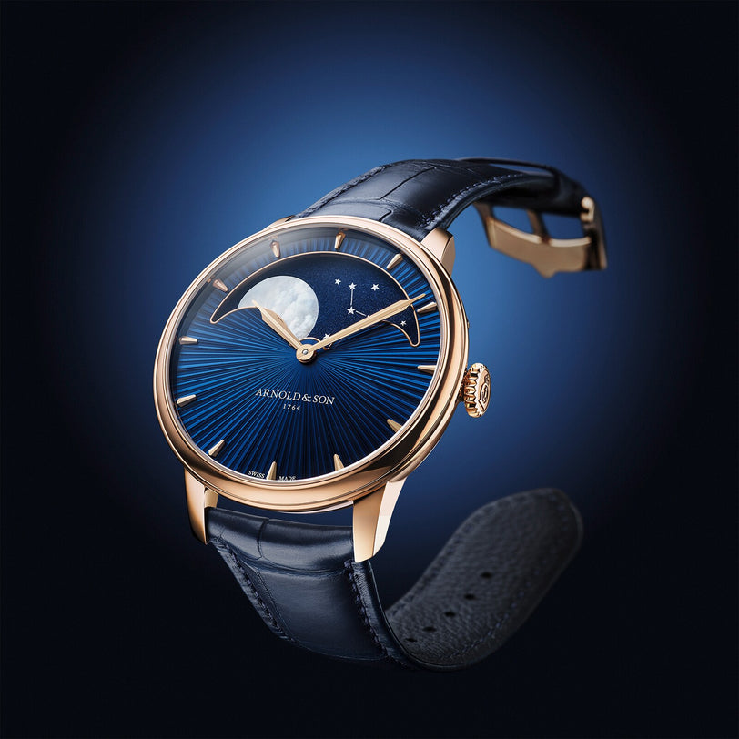 The Arnold & Son Perpetual Moon 41.5mm Red Gold features a blue dial, moonphase display, gold case, and black leather strap, showcased against a dark background.