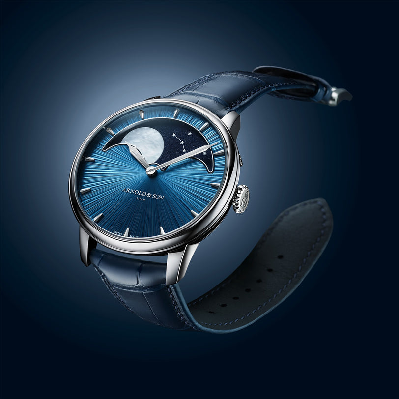 The Arnold & Son Perpetual Moon 41.5mm Platinum features a luxury blue dial with moonphase design and leather strap, showcased against a dark gradient background.