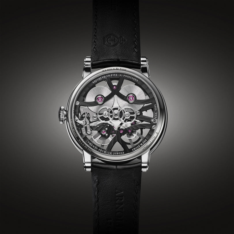 The Arnold & Son Nebula 38 Steel features visible gears and jewels in a skeleton design, a black leather strap, and a metallic case, showcased against a dark background.