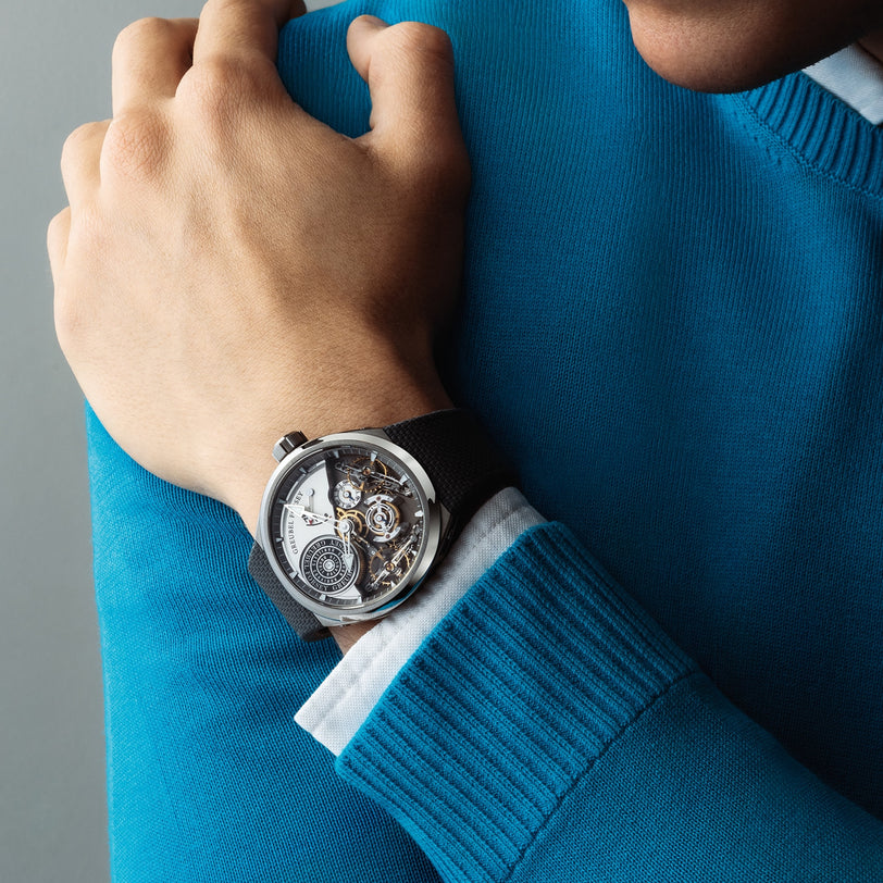 Close-up of a person wearing a blue sweater and a Greubel Forsey Double Balancier Convexe watch with exposed mechanical gears on their wrist.