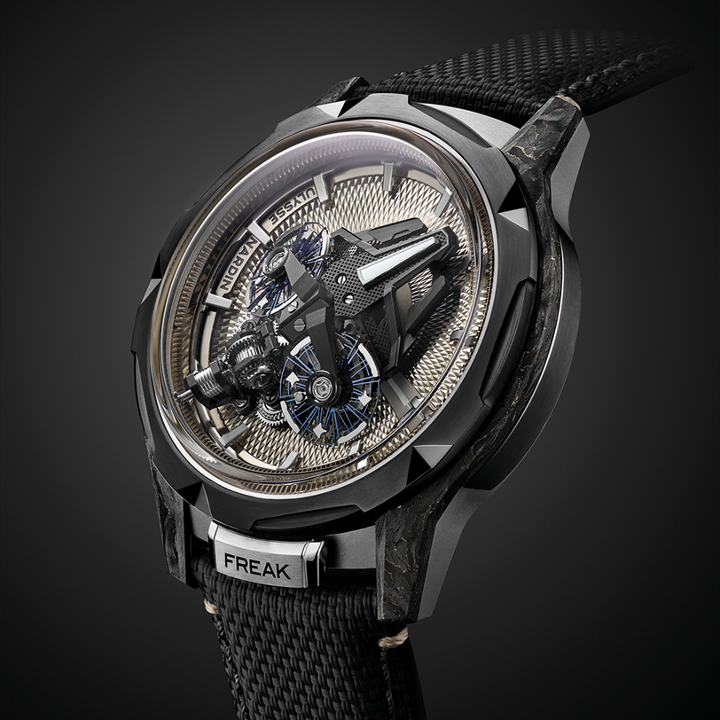 A close-up of the Ulysse Nardin Freak X Crystalium wristwatch with a black textured strap, metallic case, and visible mechanical components on the open dial. "FREAK" is engraved at the 6 o’clock position.