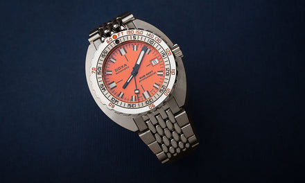 A close up view of the DOXA Sub 300T watch with a coral red-orange dial and silver-tone strap over a blue background
