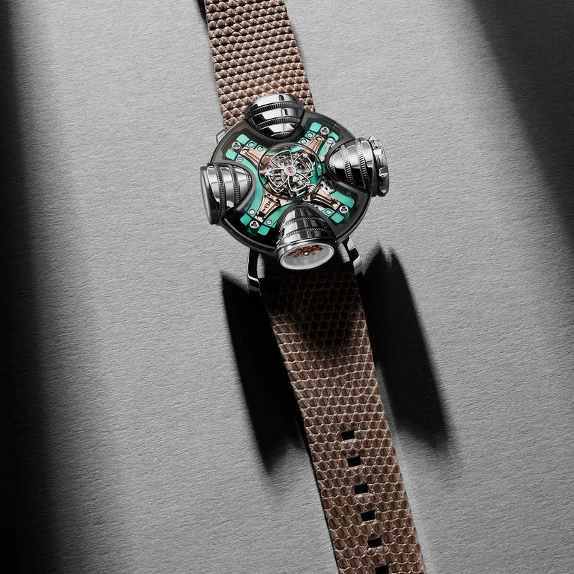 The MB&F HM11 Art Deco is a futuristic watch with a metallic, skeletonized multi-dial face showcasing exposed gears and green circuit-like details, paired with a brown textured strap, set against a gray surface with dramatic shadows.