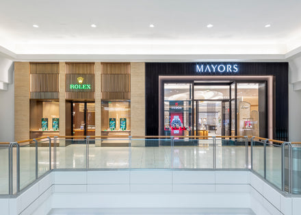 Storefronts of Rolex and Mayors jewelry stores in a shopping mall, with glass displays and bright lighting.