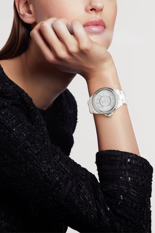 A person in a textured black top rests their chin on their hand, showcasing the Chanel J12 Diamond Bezel—a white wristwatch with a durable ceramic and steel case and a round, crystal-studded bezel—against a plain, light background.