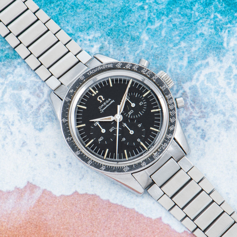 An OMEGA Speedmaster stainless steel watch with a black dial set against a beach-themed background.