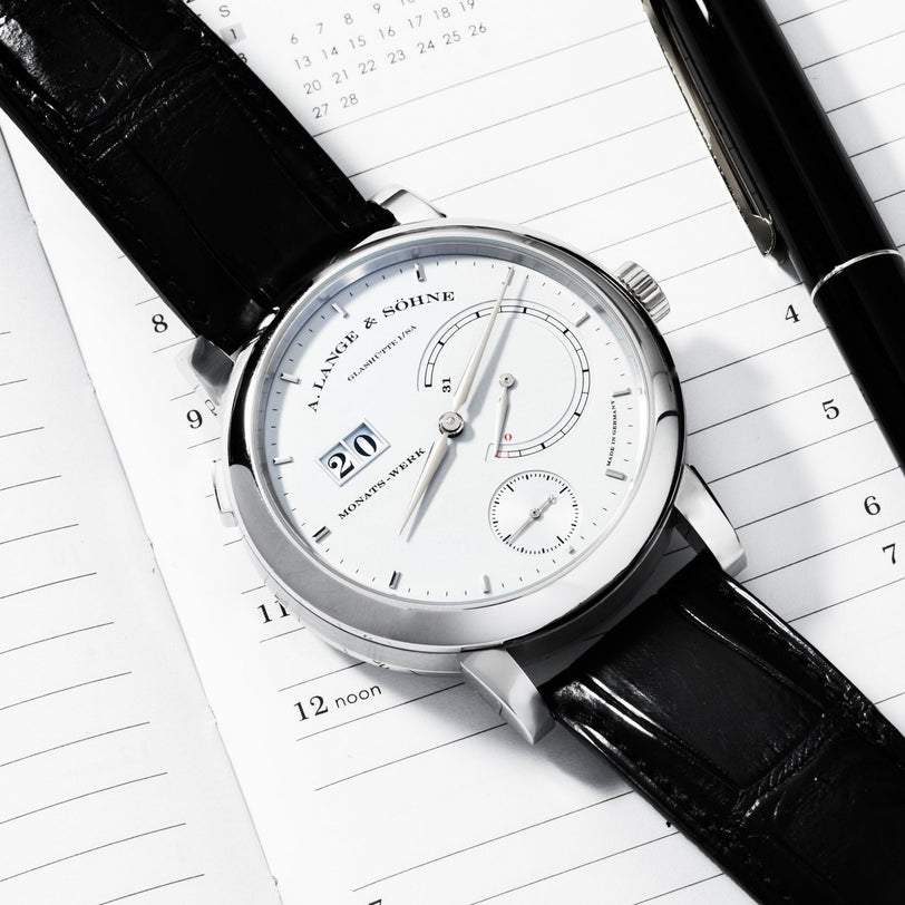 The Lange & Sohne Lange 31 luxury wristwatch with a black leather strap sits on an open planner next to a black pen.