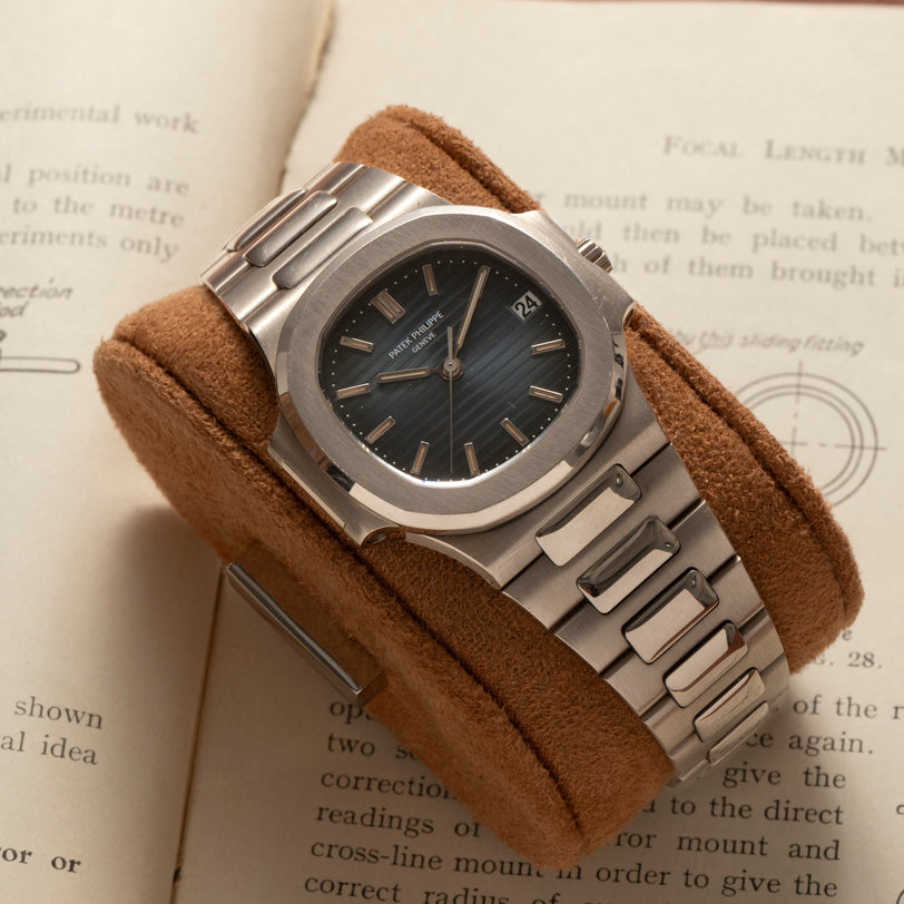 A Patek Philippe Nautilus silver luxury wristwatch with a black dial is displayed on a brown suede cushion over an open book.