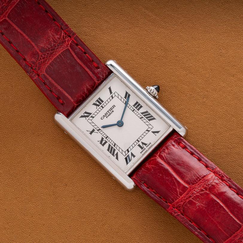 The Cartier Tank Louis is a rectangular watch featuring Roman numerals and a red leather strap on a tan surface.