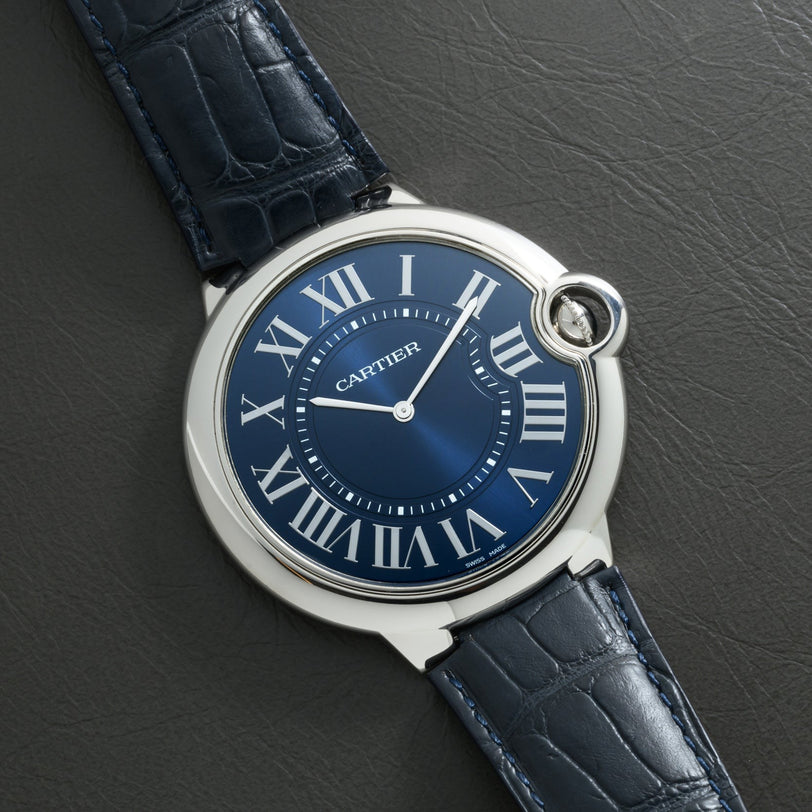 A Cartier Ballon Bleu watch with Roman numerals and a leather strap rests on a textured dark surface.