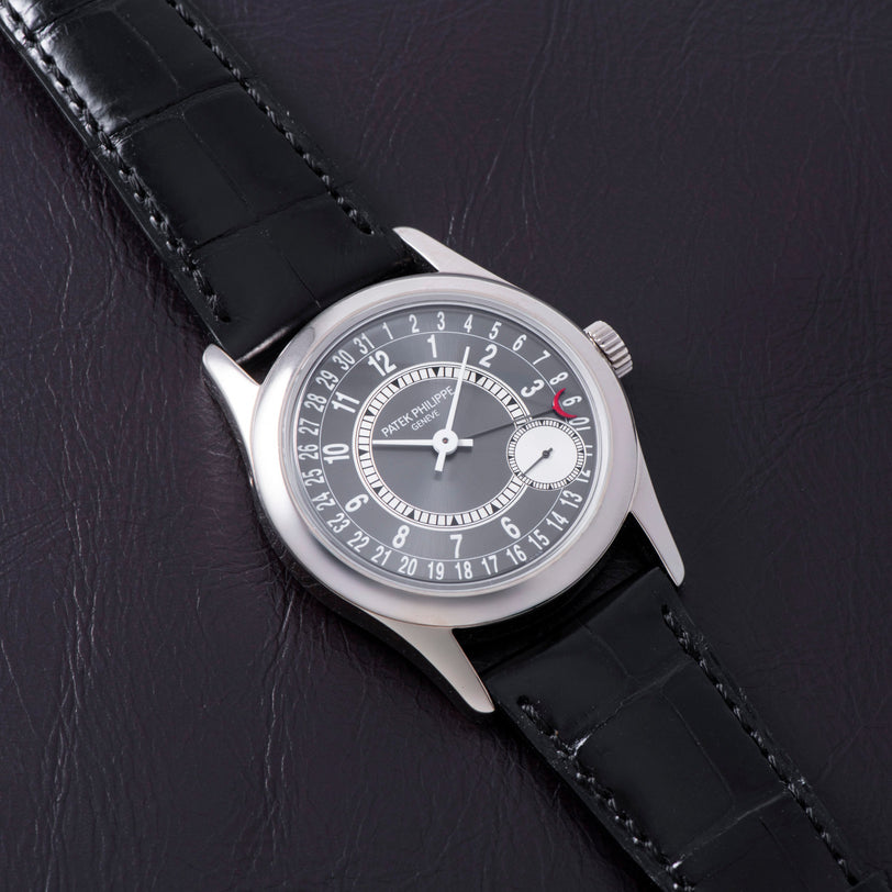 A Patek Philippe Calatrava wristwatch in silver with a black leather strap, displayed on a dark leather surface.