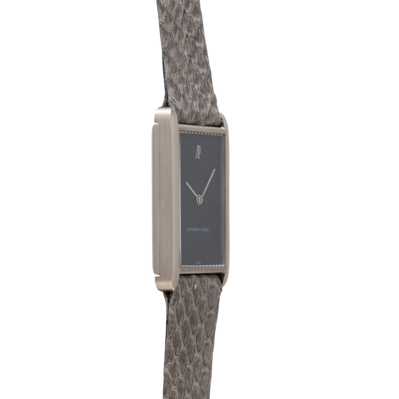 Rectangular stainless steel watch with dark dial, minimalist hands, and gray patterned fabric strap.