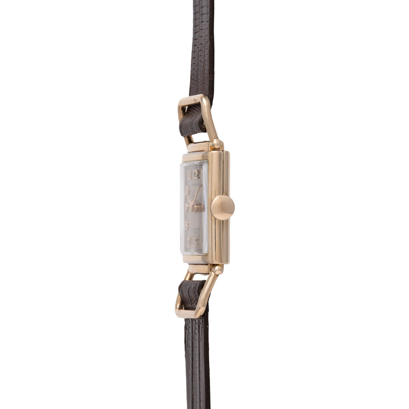 Gold-tone rectangular watch with dark leather strap and crown button on side.