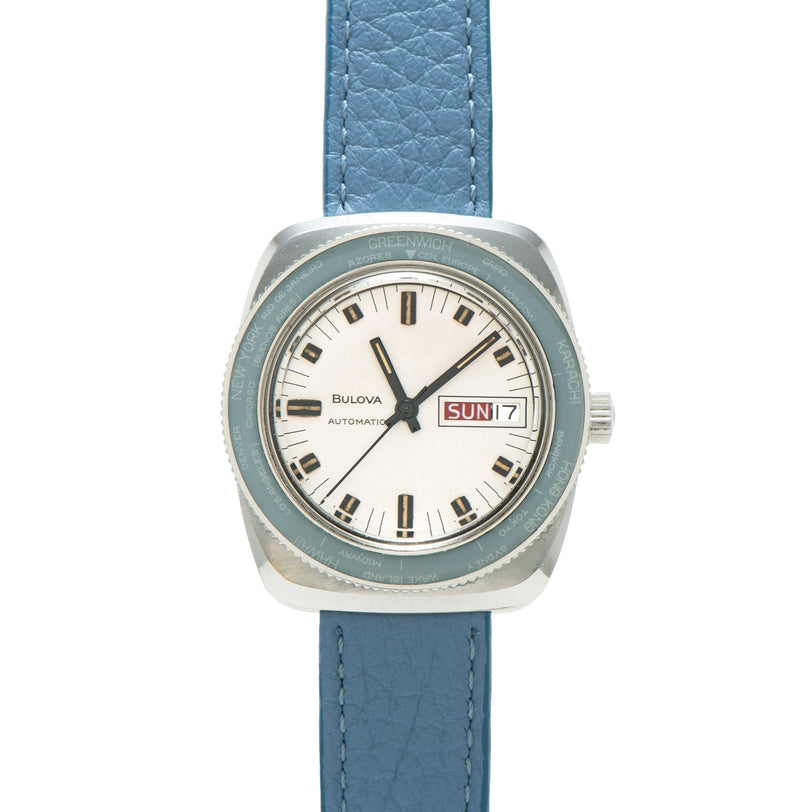 Silver-tone Bulova automatic watch with cream dial, day-date window, and blue leather strap.