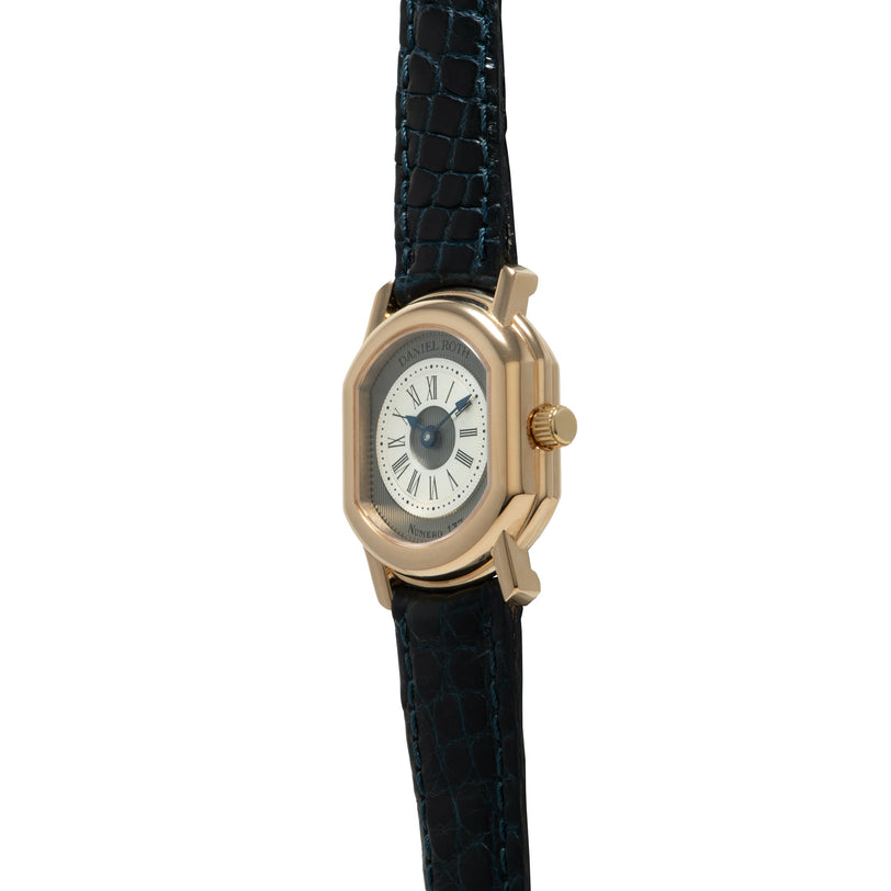 Gold-tone rectangular watch with cream dial, Roman numerals, and dark blue crocodile leather strap.