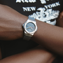 A person crosses their arms over a black tee with white New York graphics, wearing a Hublot Classic Fusion Titanium Limited Edition For Hodinkee wristwatch with a round face and gray strap.