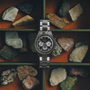 The Zenith Chronomaster Original Triple Calendar Limited Edition For Hodinkee wristwatch is showcased among colorful rocks in a segmented box set against an orange background.
