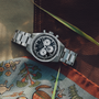 The Zenith Chronomaster Original Triple Calendar Limited Edition For Hodinkee, a stainless steel watch with a black and white dial and modern chronograph subdials, rests on patterned fabric and leather with green grass accents.