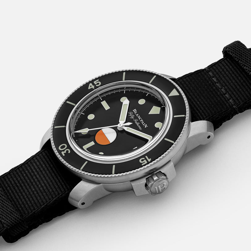 A close-up of the Blancpain Fifty Fathoms MIL-SPEC Limited Edition For HODINKEE dive watch, featuring a black fabric strap, black dial, luminous markers, and an orange-and-white segment on the lower half of the face.